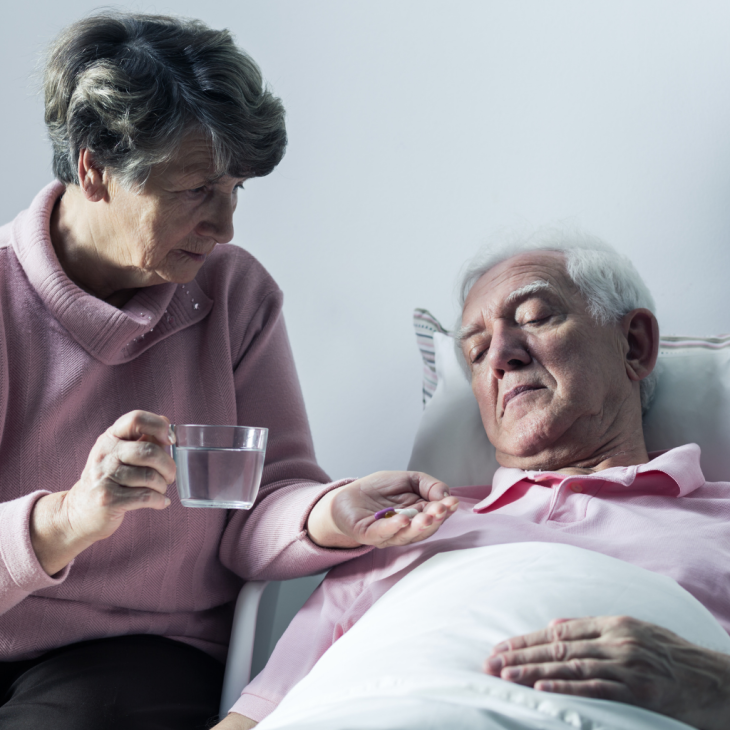 Seniorka podająca lek leżącemu seniorowi. Seniorka trzyma w ręce szklankę z wodą.