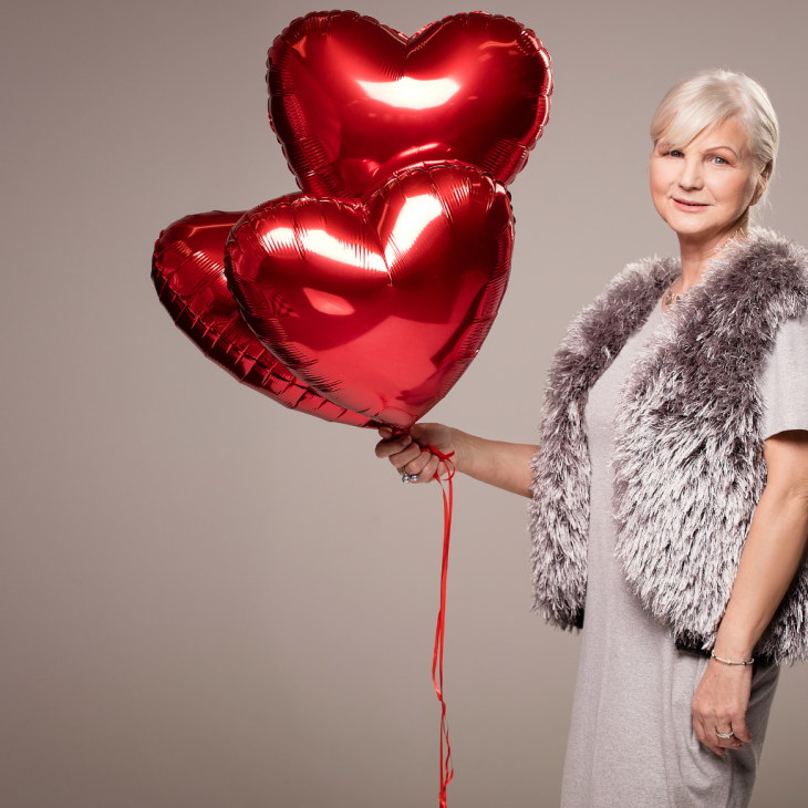 Seniorka trzyma w ręku trzy czerwone balony w kształcie serca.