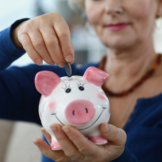 Świnka skarbonka z różowym nosem i uszami. Seniorka trzyma rękę z monetą nad skarbonką.