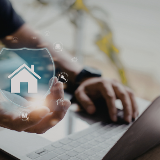 Dłonie mężczyzny, który trzyma ikonkę przedstawiającą dom. Po prawej srebrny laptop na stole.